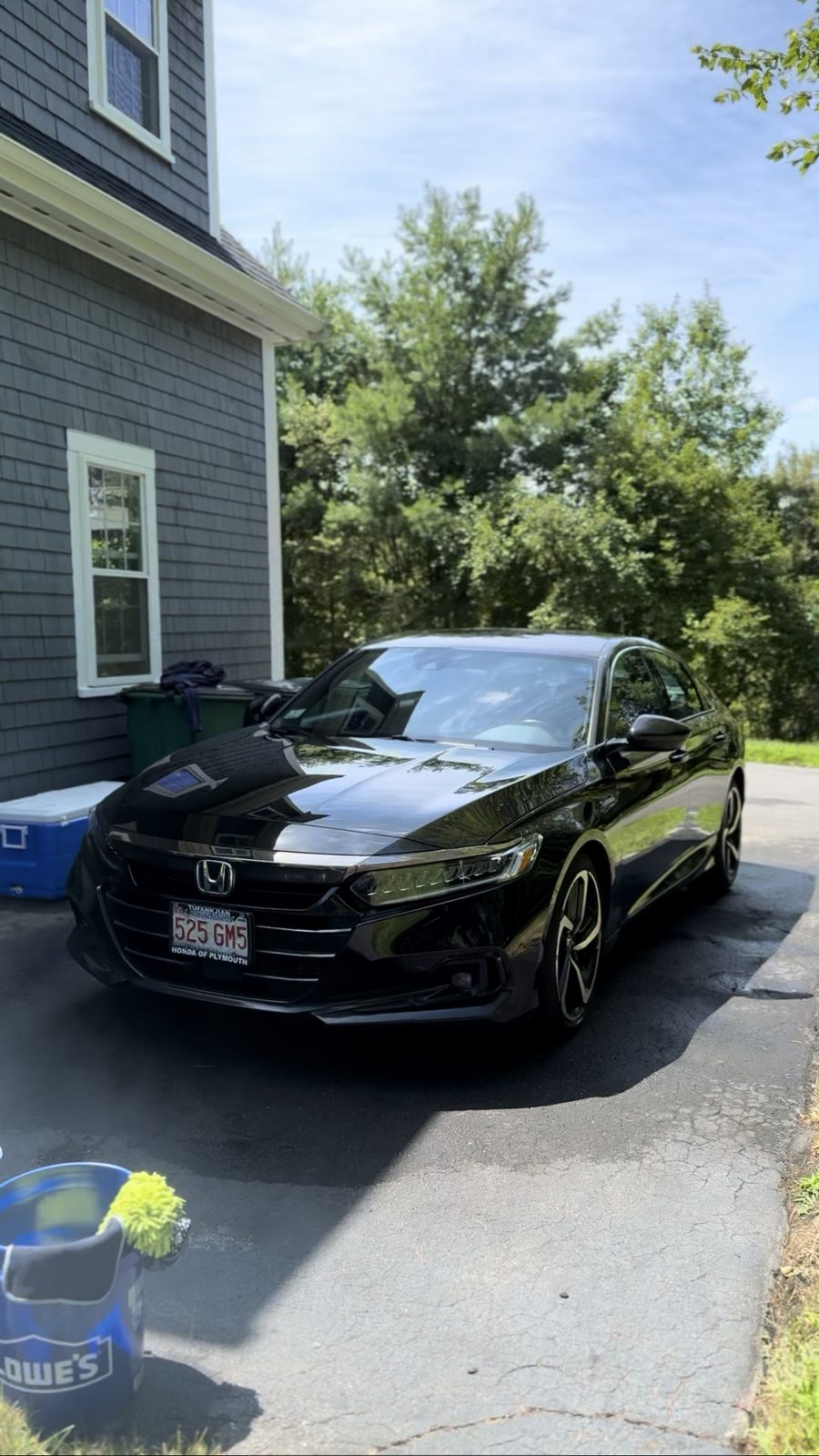 Black Honda Accord front three-quarter view with deep glossy paint reflecting trees and sky, wash bucket in the foreground