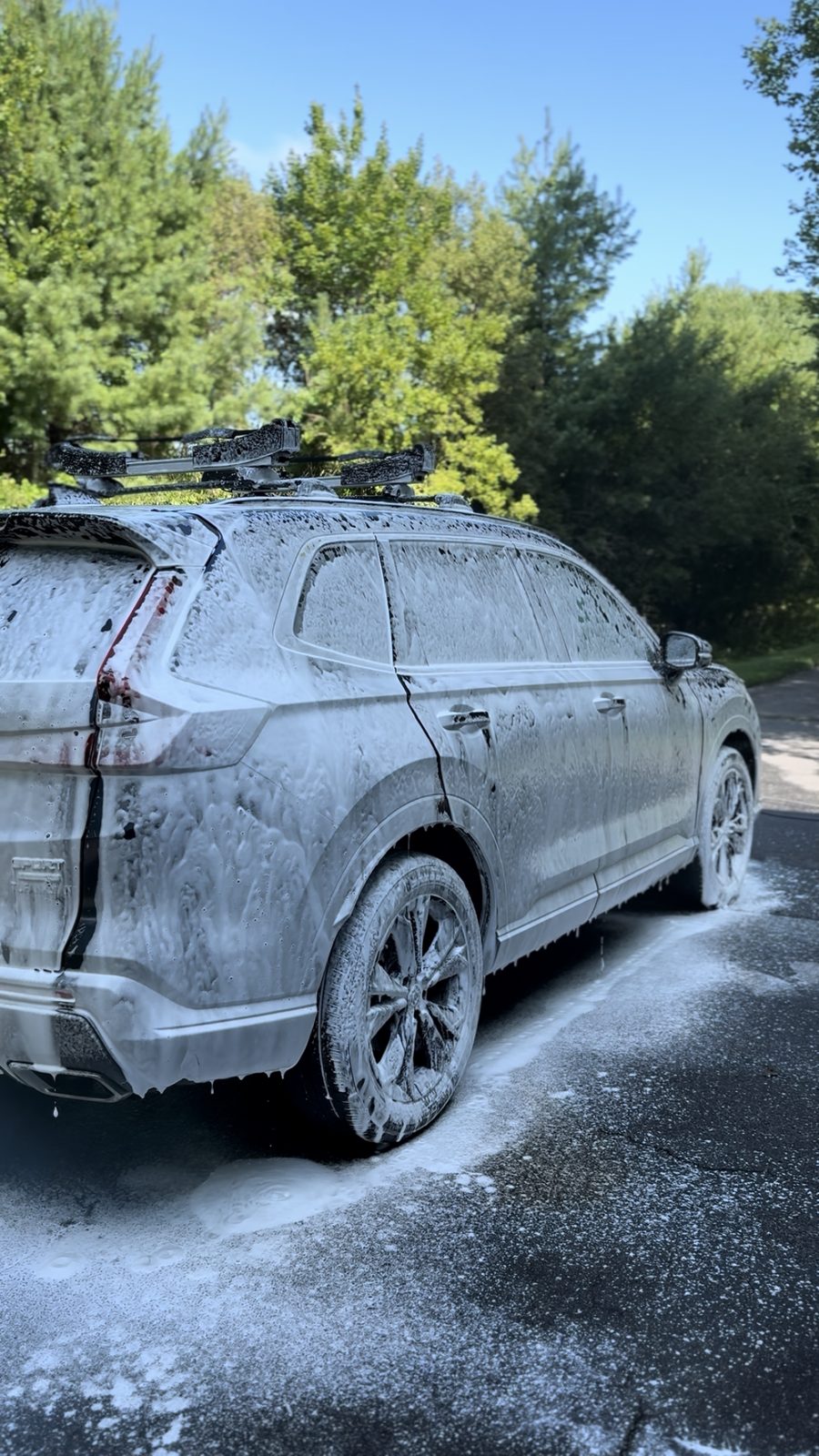 Honda CR-V with roof rack completely covered in thick white foam during a mobile foam pre-wash, suds running off onto the pavement